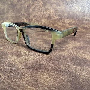 Può includere: Occhiali rettangolari con montatura bicolore. Le montature sono un mix di marrone chiaro e scuro, con un effetto sfumato. Le lenti sono trasparenti. Gli occhiali sono appoggiati su uno sfondo marrone strutturato.