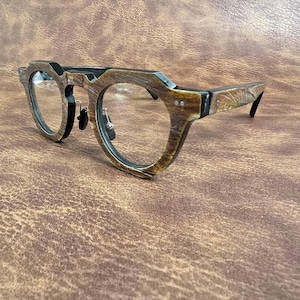 Könnte beinhalten: Brille mit einem markanten Holzrahmen und einer strukturierten Oberfläche. Die Fassungen sind eine Mischung aus Braun und Schwarz, mit runden Gläsern. Die Brille hat einen klassischen, aber modernen Stil.