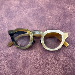 以下が含まれることがあります： ユニークな角張ったフレームデザインの眼鏡。フレームは、濃い茶色から明るいタン色へと変化する色のグラデーションが特徴です。レンズは透明で、テンプルは明るい茶色です。