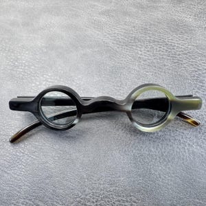 May include: Eyeglasses featuring circular frames with a smooth, polished finish in a marbled gray and black pattern. The bridge is curved and connects the two round lenses, while the temples extend outward from the sides of the circular rims.