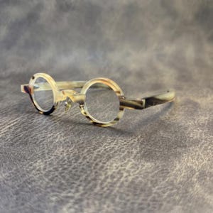 Pode incluir: Óculos redondos com armação castanha clara e creme. Os óculos têm um design clássico e vintage com ponte de fechadura e lentes transparentes. A armação apresenta um padrão marmoreado, adicionando um toque único.