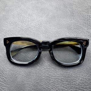 May include: Black and grey marbled acetate spectacles featuring a thick, rounded frame design. Each temple hinge is accented with three small, circular gold-toned metal rivets on the front corners, providing a distinct and structured appearance.