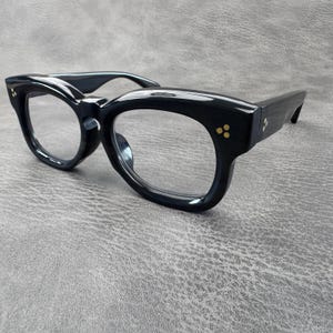 May include: Glossy black thick-rimmed glasses featuring clear lenses and a bold frame design. The front corners and the temples are accented with small, gold-coloured three-dot rivets, providing a distinct detail to the solid black frame structure.