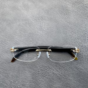 Può includere: Occhiali senza montatura con lenti trasparenti, un ponte in metallo dorato e cerniere in metallo dorato collegate ad aste nere. Il design della montatura include una sottile barra superiore dorata e naselli trasparenti fissati al ponte.