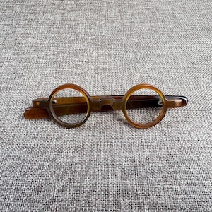 Könnte beinhalten: Runde, braune Brille mit Schlüssellochsteg und dunkelbraunem Rahmen. Die Brille hat ein klassisches, Vintage-Design. Die Gläser sind klar, und der Rahmen besteht aus einem durchscheinenden Material. Die Brille liegt auf einer strukturierten, neutralfarbenen Oberfläche.