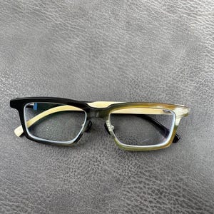 May include: Rectangular eyeglasses with a black and olive green frame and light-colored temples. The lenses are clear, and the glasses are set against a gray background. The glasses have a modern, stylish design.