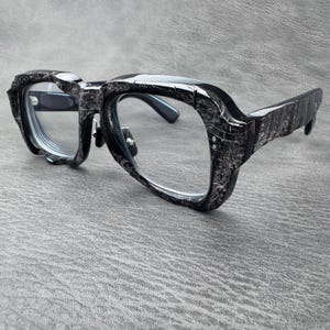 May include: Spectacles featuring thick, chunky frames with a textured, cracked black and grey finish. The glasses have clear lenses, prominent bridge detailing, and sturdy temples with visible metal rivets on the front corners of the frame.