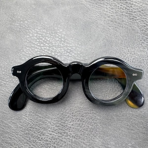 May include: Round spectacles featuring a thick, polished black frame with a subtle horn-like pattern visible on the inner edges. The glasses have two small silver-toned rivets on each side of the front frame and curved temples extending outwards.
