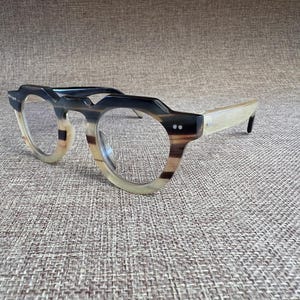 May include: Eyeglasses with a unique design. The frames feature a combination of black, brown, and cream colors. The lenses are clear, and the overall shape is round with a distinctive angular top. The temples are a light cream color.