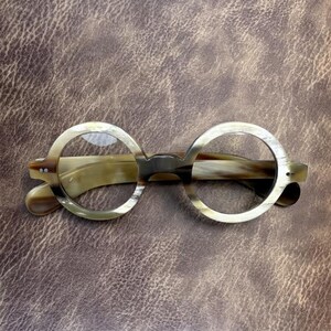 May include: Round-framed spectacles with a light brown and cream marbled pattern. The glasses have a keyhole bridge and a classic, vintage aesthetic. The frames are set against a textured brown background.