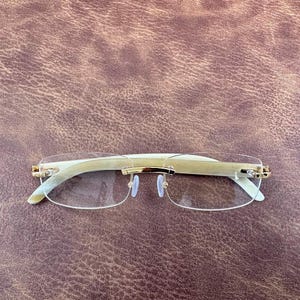 Può includere: Occhiali senza montatura con montatura colorata chiara e dettagli dorati. Gli occhiali hanno una forma rettangolare e lenti trasparenti. Le aste sono di colore beige chiaro. Gli occhiali sono su uno sfondo marrone.