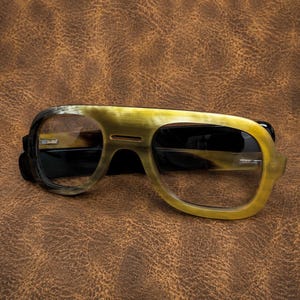 May include: Aviator-style spectacles with a distinctive horn-rimmed design. The frames display a gradient of colours, shifting from dark brown to a light yellow-brown. The lenses are clear, and the temples are black. The spectacles rest on a textured brown surface.