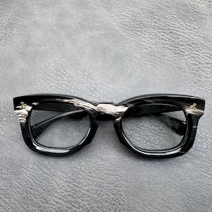 May include: Spectacles featuring a thick, black and white marbled acetate frame with a distinct wood-grain pattern. The front of the frame includes small, gold-toned metal rivets at each outer corner, and the lenses are clear and transparent.