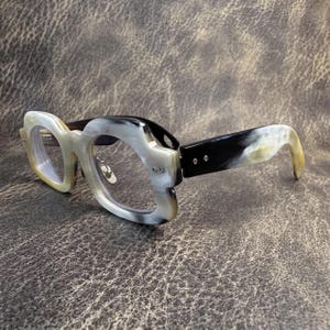 May include: Eyeglasses with a distinctive design. The frames are a mix of cream, black, and tan colours. The arms are black with cream and tan tips. The lenses are clear.