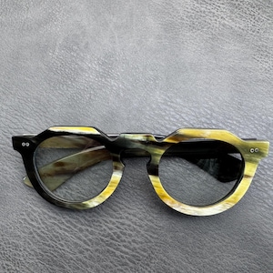 Puede incluir: Gafas con una montura gruesa y redondeada que presenta un patrón jaspeado en tonos negros, amarillos y crema. La montura tiene un borde superior angular distintivo y dos pequeños remaches de color plateado a cada lado del frente.