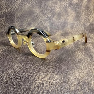 May include: Round-framed spectacles with a distinctive marbled design in tan, cream, and black. The glasses feature a thick frame and a classic, vintage style. The lenses are clear, and the arms are slightly curved.
