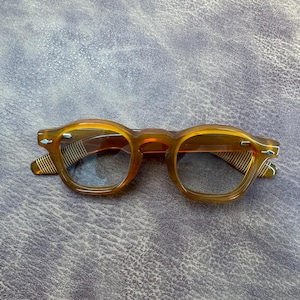 Puede incluir: Un par de gafas marrones con montura rectangular redondeada. Las gafas tienen un tinte marrón claro y un patrón de rayas en las patillas. Las lentes son transparentes.