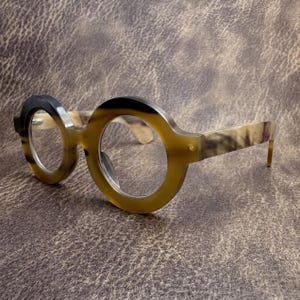 May include: Round-framed spectacles with a distinctive design. The frames are a mix of light brown and black, with a marbled pattern on the arms. The lenses are clear, and the glasses are set against a textured brown background.