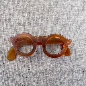 Peut inclure: Lunettes à monture ronde de couleur marron chaud. Les lunettes présentent une monture et des branches épaisses et arrondies, avec un aspect légèrement translucide. Les verres sont clairs et le design général est d'inspiration vintage.
