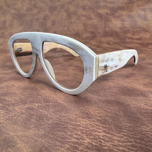 May include: Spectacles with a thick, light-coloured frame featuring a natural, marbled grain pattern. The frame has a distinct, wide bridge design connecting the two large, rounded lenses. The temples are wide and match the frame's marbled texture.