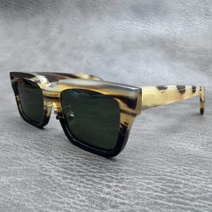 Puede incluir: Gafas de sol con una montura rectangular gruesa con un patrón jaspeado similar al cuerno en tonos crema, bronceado y marrón oscuro. Las lentes son de color verde oscuro y las patillas son anchas, a juego con el patrón orgánico y variado.