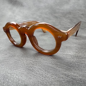 May include: A pair of thick-rimmed spectacles featuring a translucent amber-brown frame with a distinct rounded shape. The frames have a smooth, polished finish and include two small silver-toned metal rivets on each side of the front frame.