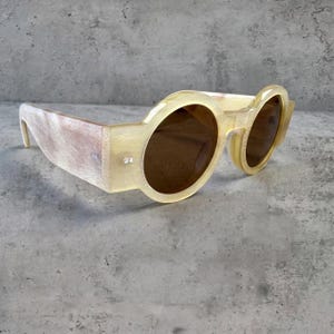 May include: Unique, oversized sunglasses with a light yellow frame and brown lenses. The frame has a rectangular shape with rounded lenses. The arms of the sunglasses have a light brown, textured pattern. The sunglasses are a bold fashion statement.