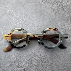 May include: Round eyeglasses featuring a mottled horn-like pattern in shades of brown, gray, and cream. The frames have a distinct bridge connecting the circular rims, with integrated nose pads and temples extending from the sides of the frame front.