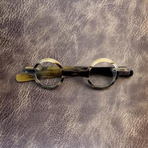 May include: Circular-framed spectacles with a marbled effect in brown, tan, and black. The glasses have a vintage design, with a keyhole bridge and curved arms. The lenses are clear, and the frame appears to be made of a durable material.