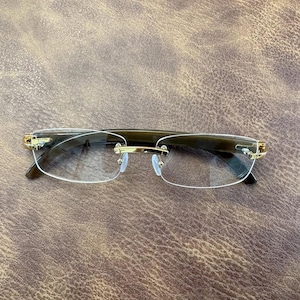 May include: Rimless spectacles with a dark brown frame and gold-coloured accents. The lenses are clear, and the spectacles have a rectangular shape. The spectacles are set against a brown leather-textured background.