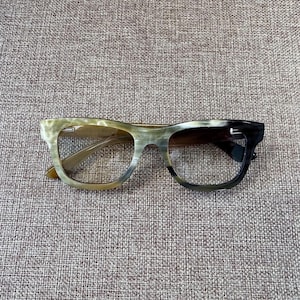 Puede incluir: Gafas con un diseño de montura único. Las monturas hacen una transición de verde claro a marrón oscuro y negro. Las gafas tienen forma rectangular y están sobre un fondo neutro.