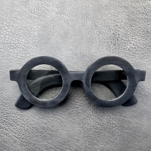 May include: A pair of round-framed glasses with a matte black finish. The frames feature a thick, circular design around the lenses with a straight bridge connecting the two sides and matching black temples extending from the outer edges.