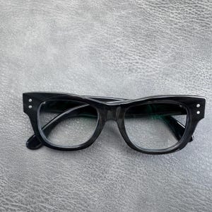 May include: A pair of black eyeglasses with a thick, glossy frame and a distinct cat-eye shape. The front of the frame features two small silver-toned metal rivets on each outer corner, and the lenses are clear and transparent.