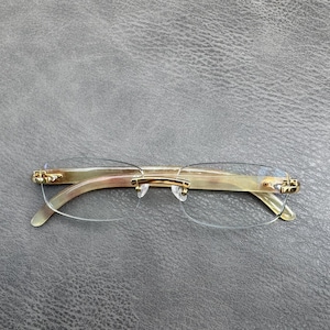 Pode incluir: Óculos sem aro com lentes transparentes e uma ponte de metal em tom dourado. As hastes são feitas de um material marmorizado em bege claro e castanho, ligadas às lentes com dobradiças e componentes de metal em tom dourado.