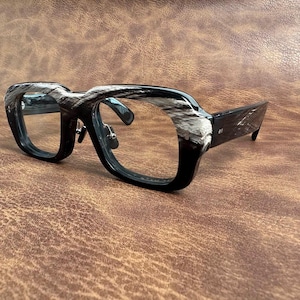 May include: Black and white patterned eyeglasses with a thick frame. The glasses have a bold, angular design with a marbled effect on the upper frame. The lenses are clear, and the temples are solid black.