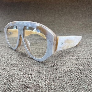 May include: Oversized spectacles featuring a thick, polished frame with a marbled pattern in shades of cream, beige, and light grey. The glasses have clear lenses and a distinctive bridge design that connects the two large, rounded frames.