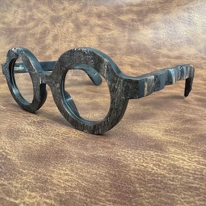 May include: Round-framed spectacles with a dark, textured finish. The frames have a unique, organic appearance, with visible grain and a mottled colour pattern. The lenses are clear, and the arms curve gently for a comfortable fit.