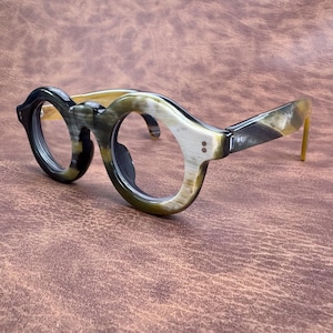 May include: Eyeglasses with a distinctive horn-rimmed design. The frames combine dark green, black, and tan tones, with a rounded shape. The temples are a lighter tan colour. The glasses are set against a brown background.