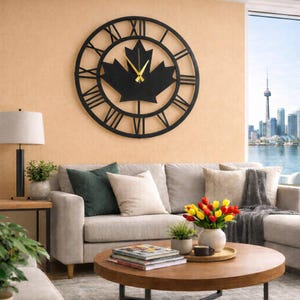 Peut inclure: Grande horloge murale en métal noir avec un motif de feuille d'érable au centre. L'horloge affiche des chiffres romains et des aiguilles dorées. Elle est fixée sur un mur beige au-dessus d'un canapé gris.
