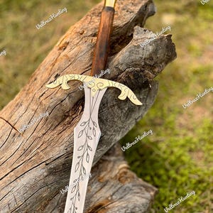 May include: A decorative sword with a wooden handle and a gold-colored hilt. The blade is silver with a leaf pattern. The sword is resting on a piece of wood. The text "Veltraz Hunting" is visible in the image.