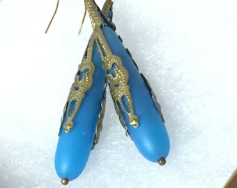 May include: A pair of blue teardrop earrings with gold-colored filigree accents. The earrings have a matte finish and are suspended from gold-colored ear wires. The earrings are displayed in a white box.