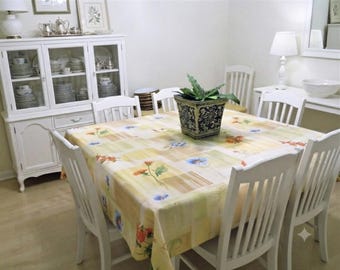 Puede incluir: Una mesa de comedor cuadrada con un mantel amarillo claro con estampado floral, rodeada por cuatro sillas de madera blanca. Una maceta cuadrada con estampado en blanco y negro con una planta verde se sitúa en el centro de la mesa.