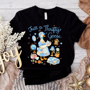 Puede incluir: Camiseta negra con la frase "Just a Thrifty Goose" en escritura azul claro. El gráfico presenta un ganso con gorro y delantal, rodeado de objetos vintage como un globo terráqueo, un reloj y tazas de té. Un diseño encantador y caprichoso.