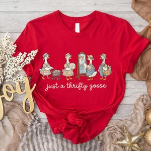 Puede incluir: Camiseta roja con una ilustración de dibujos animados de gansos vestidos con trajes vintage, comprando y mirándose en un espejo. El texto "just a thrifty goose" está escrito en una fuente de guión en la parte delantera.