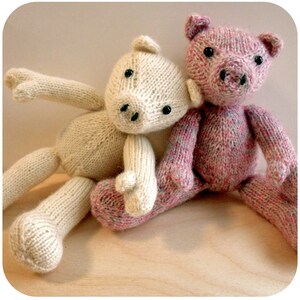 Könnte beinhalten: Zwei gestrickte Stofftiere, eines weiß und eines rosa, sitzen zusammen. Das weiße Tier hat eine lange Schnauze und schwarze Augen, während das rosa Tier eine kürzere Schnauze und eine rosa Nase hat.