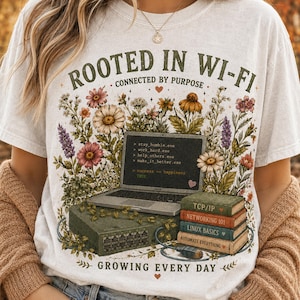 May include: A white t-shirt featuring a graphic of a laptop and a stack of books surrounded by colorful wildflowers. The text reads ROOTED IN WI-FI, CONNECTED BY PURPOSE, and GROWING EVERY DAY. The book spines are labeled TCP/IP, NETWORKING 101, LINUX BASICS, and AUTOMATE EVERYTHING.