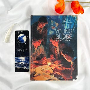 Op de afbeelding: Een boek getiteld "All The Young Dudes Book Two" met een bijpassende bladwijzer. De boekomslag toont een groep mensen in een schemerige kamer. De bladwijzer heeft een maan- en oceaanontwerp met de tekst "All the young dudes".