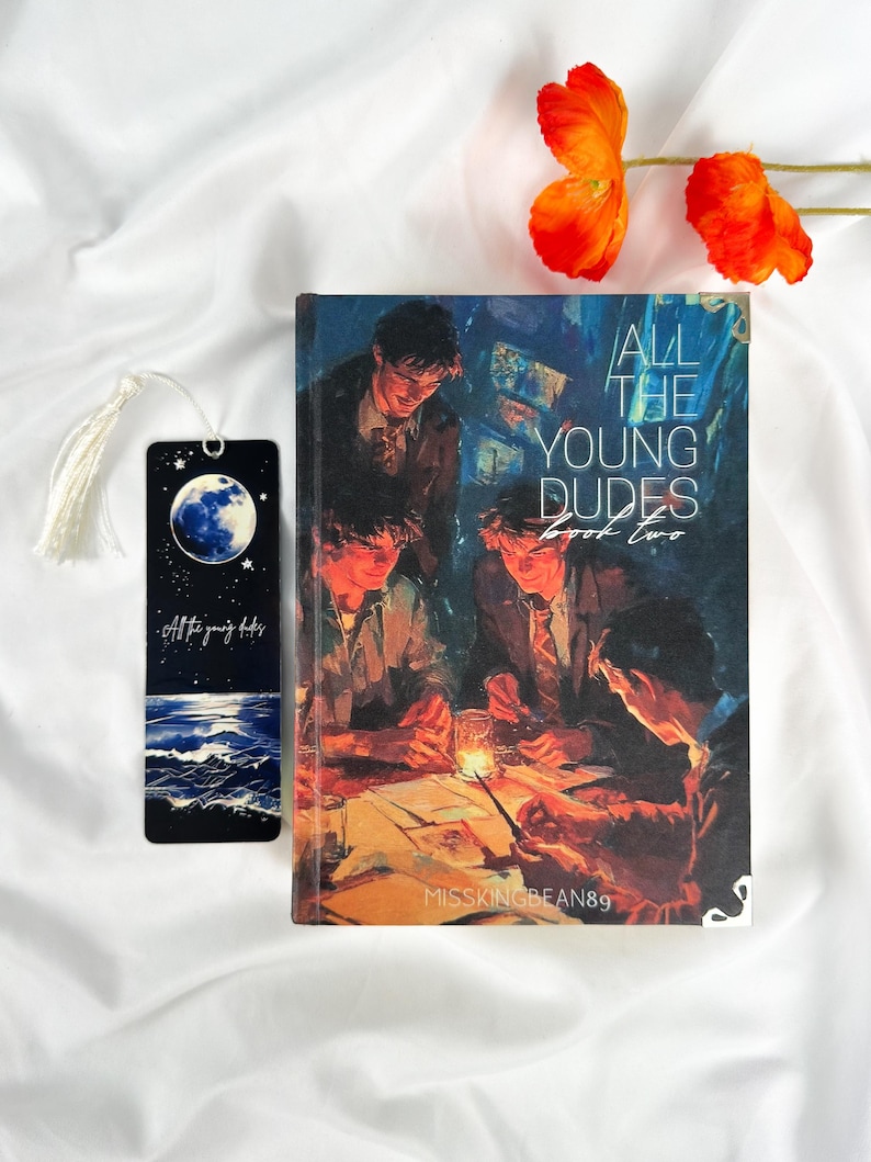 Op de afbeelding: Een boek getiteld "All The Young Dudes Book Two" met een bijpassende bladwijzer. De boekomslag toont een groep mensen in een schemerige kamer. De bladwijzer heeft een maan- en oceaanontwerp met de tekst "All the young dudes".