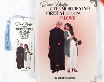 Könnte beinhalten: Ein Buch und ein Lesezeichen mit dem Titel "Draco Malfoy & The Mortifying Ordeal of Being in Love". Das Buchcover zeigt Illustrationen von zwei Charakteren. Das Lesezeichen hat eine blaue Quaste und wiederholt den Titel und das Bild des Buches.