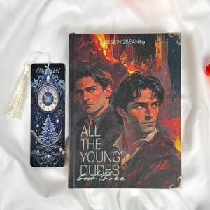 Op de afbeelding: Een boek getiteld "ALL THE YOUNG DUDES Book Three" met een omslag met twee mannen en een vurige achtergrond. Een bijpassende bladwijzer met een klokontwerp en kwast is ook aanwezig. De items staan op een witte stoffen achtergrond.